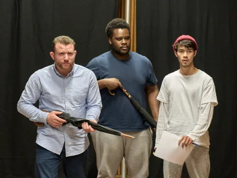 Richard James Neale (brown hair, brown beard, light blue shirt), Tomi Ogbaro (black hair, black beard, navy t-shirt), Sky Yang (black hair, black beard, red beanie), all stand looking intently at something off camera. James and Tomi hold umbrellas.