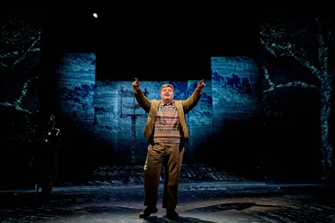 Samuel Creasey (Light brown hair, a patterned jumper and plain coat), stands centre stage with bot arms pointing at the audience.