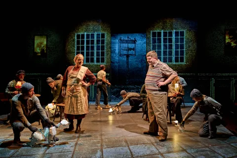 The cast of The Book of Dust on stage, there are wooden tables and chairs with cast sitting in a busy pub.
