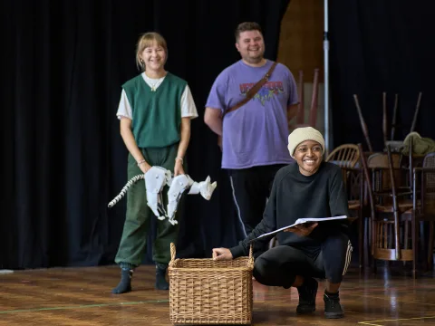 Heather Forster (blonde hair tied back, white tshirt with green sweater vest) holds origami style puppet of a cat daemon. Samuel Creasey (blonde hair, purple t-shirt), stand behind Ella Dacres (cream beanie, black jumper) who crouches next to a wicker bas