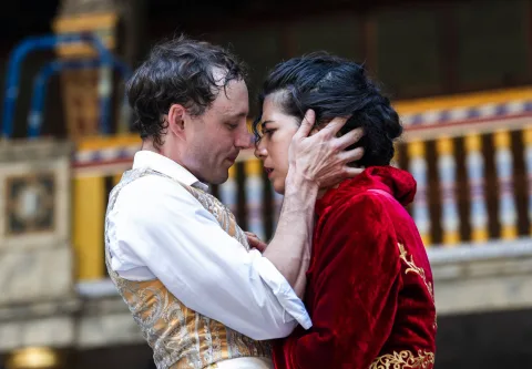 A white male actor dressed in gold and white, grasping the face of a white female actor with dark curly hair dressed in red velvet, as if in a passionate embrace.