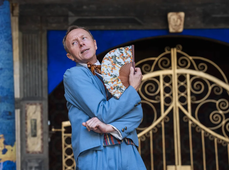 A white actor in a light blue suit poses with a floral fan.