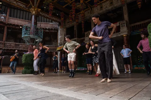 A group of actors jump in unison on a wooden stage inside a wooden theatre. They stand spaced apart with their arms on their waists.
