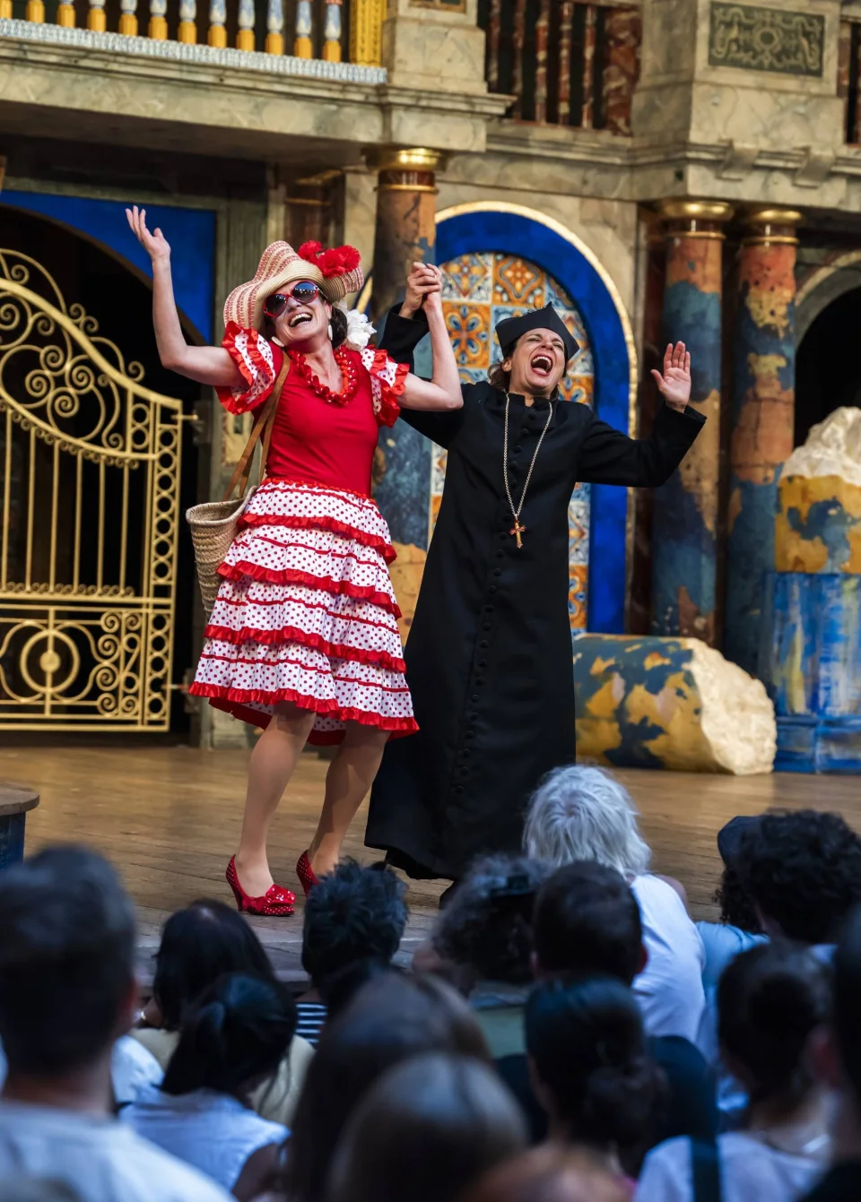 An actor in a frilled polka dot dress and an actor in a black curate's outfit embrace with their arms raised.