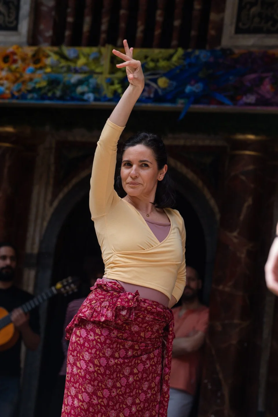 A person wearing a yellow top and a red skirt stands with one arm raised in the air and one arm on their waist.