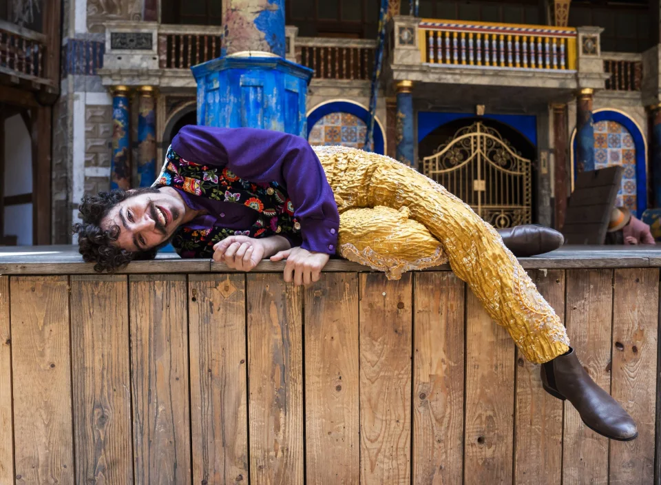 An actor with dark curly hair and a moustache lying on the stage, dressed in a purple shirt and yellow trousers.