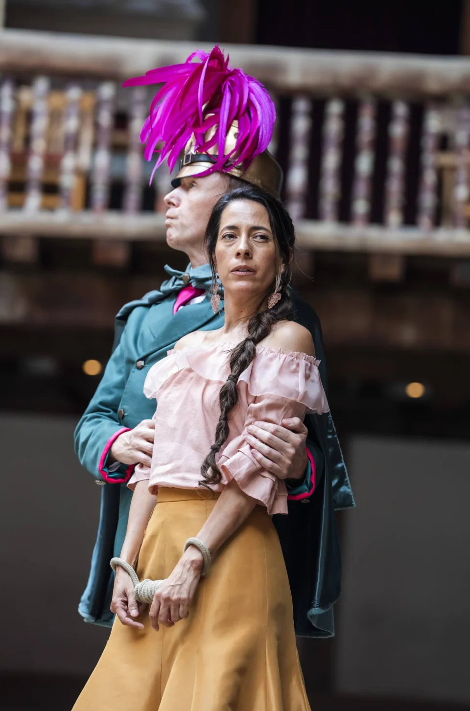 A white male actor with a pink feather plumed hat gripping the arms of a Latin American Mexican actress dressed in a pink blouse and yellow skirt.