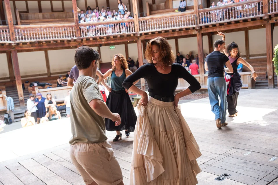 A white man wearing a green t-shirt dances on a wooden stage with a white female with dark hair who has her hands on her hips. In the background, others dance and people sit in the galleries of a wooden theatre.