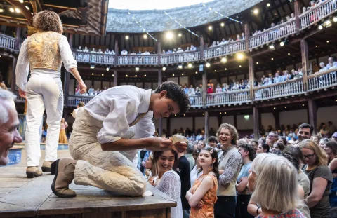 An actor dressed in white bends down into the audience from the side of the stage, clutching a scrap of paper.