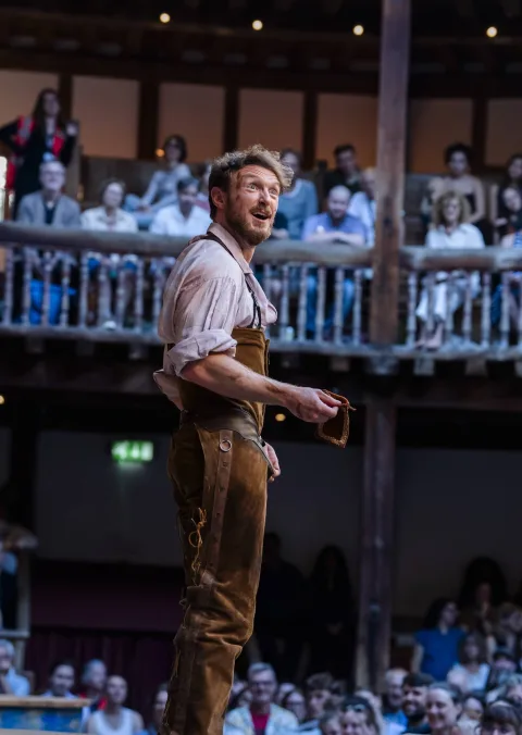 An actor in a pink shirt and brown leather trousers looks upwards, away from audience members sitting on a balcony in the background.
