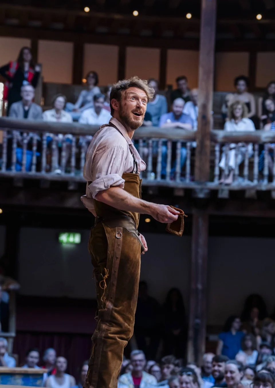 An actor in a pink shirt and brown leather trousers looks upwards, away from audience members sitting on a balcony in the background.