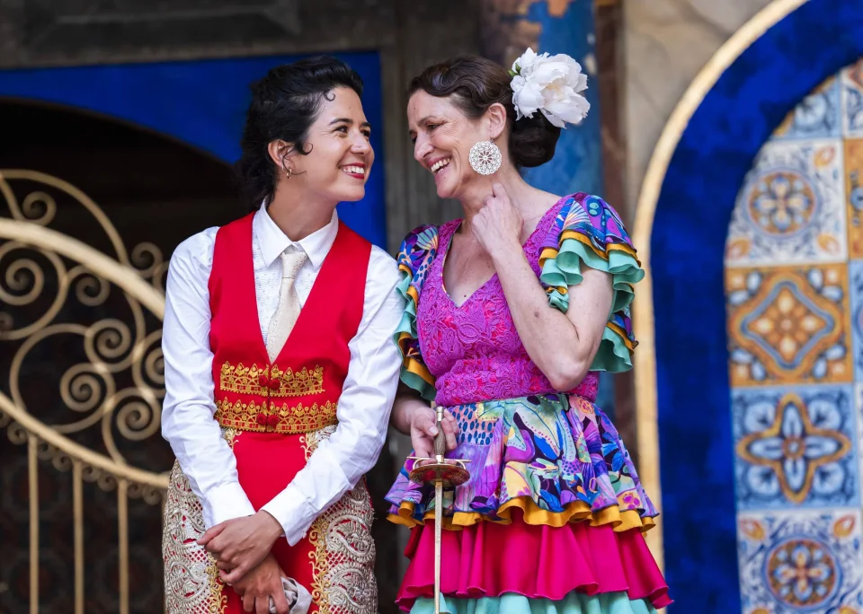 Two women smiling at one another. One is dressed in white and red, and the other in a brightly coloured flamenco dress with a white flower in her brown hair.