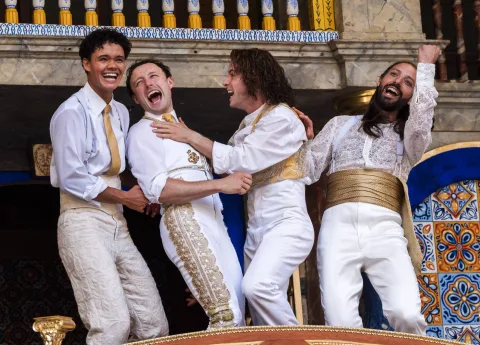 Four men dressed in gold and white, clutch each other laughing.