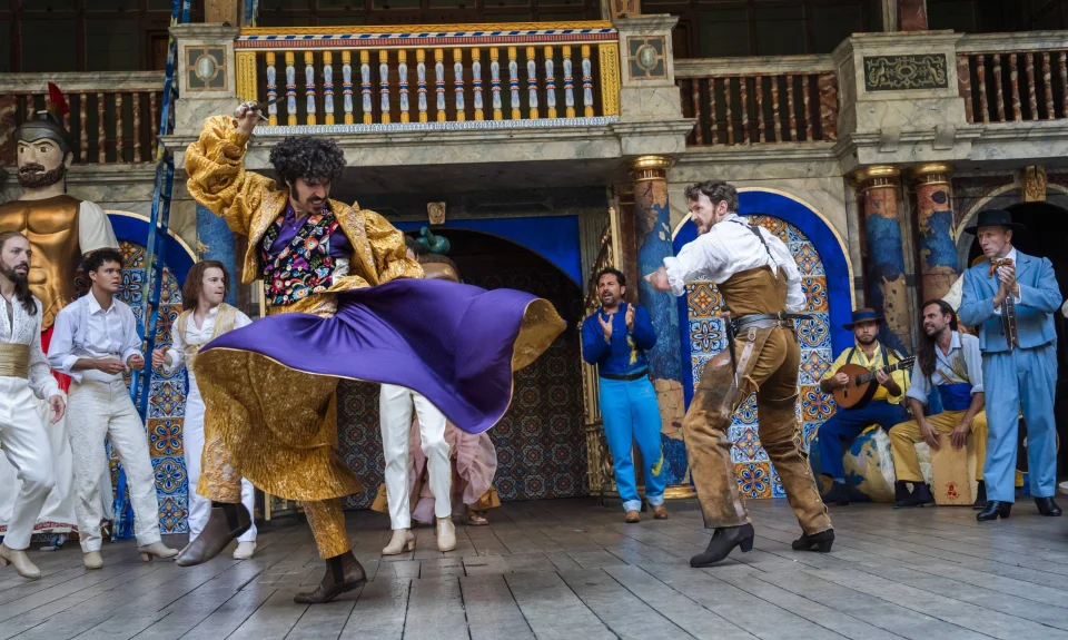 An actor twirling in a gold and purple cape, opposite an actor dressed in a white shirt and brown trousers.