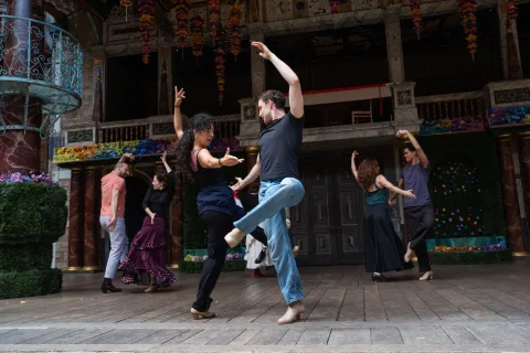 Two people dance on a wooden stage, each with one arm and one leg raised. In the background, other couples dance in the same style.