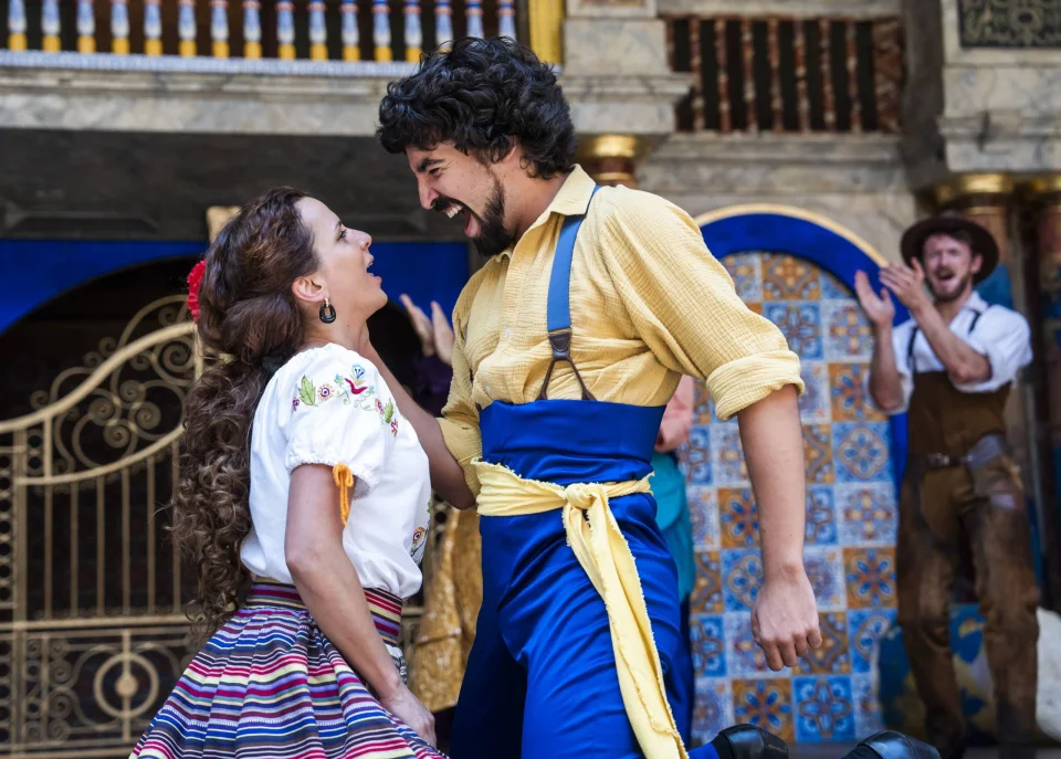 A mediterranean actor with brown curly hair in a colourful flamenco skirt and wite top, face to face with a dark-haired mediterranean dancer dressed in yellow and blue.