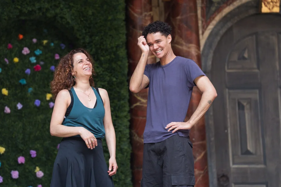 A Mediterranean/ Middle Eastern looking woman with wavy hair stands beside a Mixed Afro-Caribbean/white man with short brown curly hair. They are laughing.