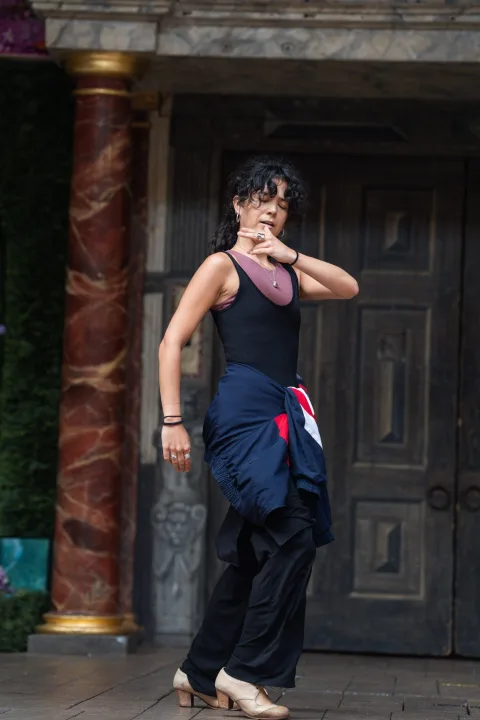A white woman with black curly hair, wearing a black vest and coat tied around her waist, dances flamenco on a wooden stage. A red marble pillar can be seen behind her.
