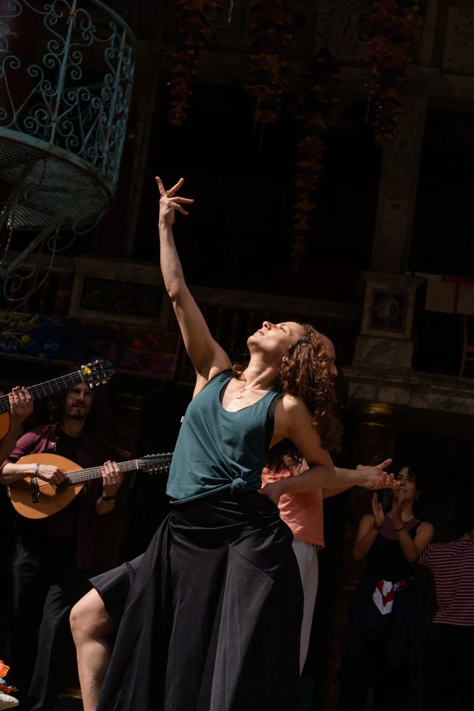 A Mediterranean/ Middle Eastern looking woman stands and leans back, with one arm raised in the air and one arm on her waist. Behind her are musicians playing instruments.