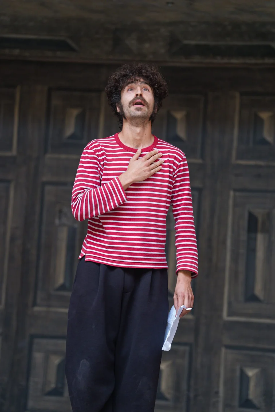 A person wearing a red and white stripy top stands with one hand on their heart and one hand holding a script, looking up above the camera.
