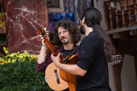 Two people stand holding guitars and facing each other. Behind them is a marble pillar with shrubbery around it's base.