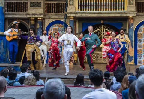 A group of actors in brightly coloured costumes dancing on the Globe stage.