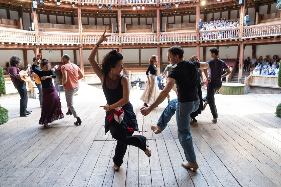 A group of people dance on a wooden stage. They are divided into partners and each make different movements. Behind them is a wooden theatre, with some groups of audience members watching.