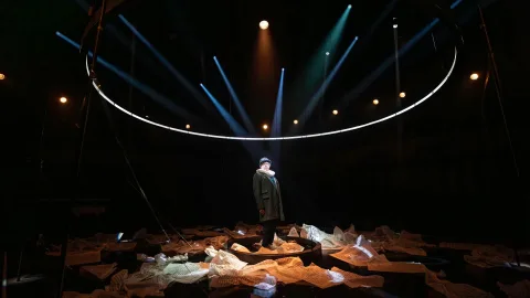 Phelim McDermott stands in the centre of the stage, surrounded by sheet music on the floor, wearing a long coast and bowler hat