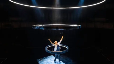 Phelim McDermott stands inside a light up ring of light, looking up to the sky with his hands in the air