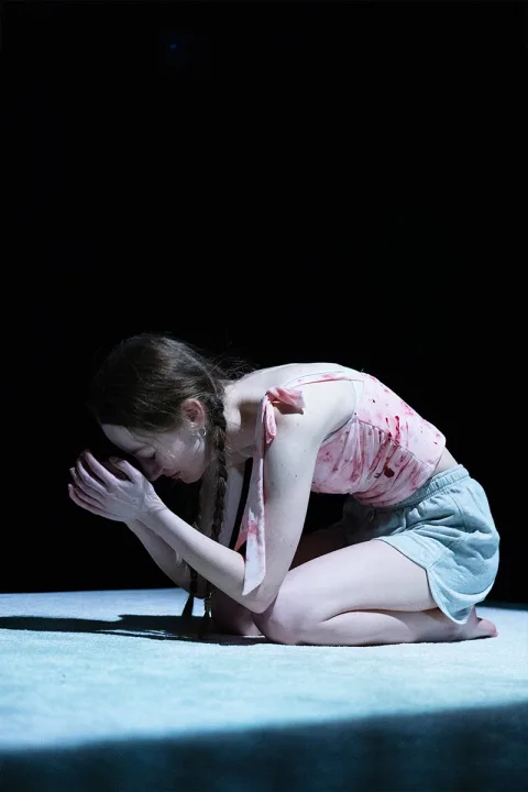Tia (Mollie Milne) kneels in a ball her hands held up to her face in a gesture of prayer.