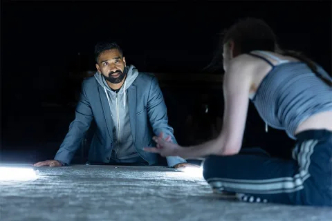 AJ (Taqi Nazeer) stands resting his hands on the grey flat surface in front of him, he is looking at Tia (Mollie Milne) who is an out of focus seated figure closer to the camera.