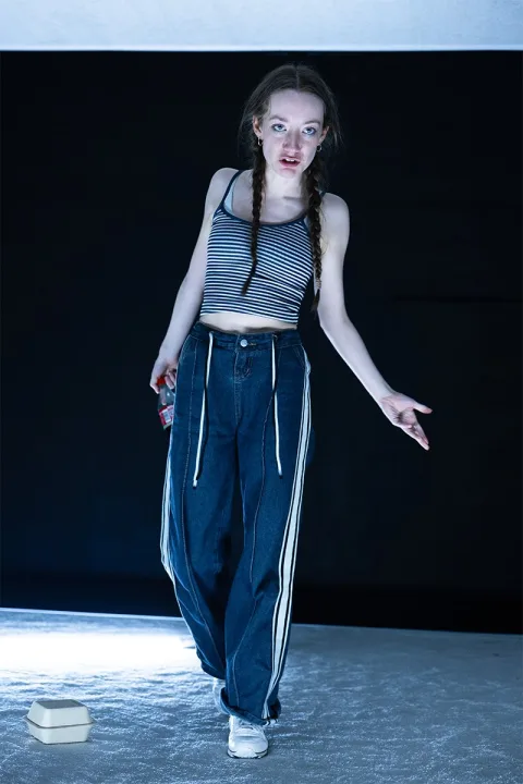 Tia (Mollie Milne) stands in a limp position, her hair in plaits and a bottle of coke in one hand.