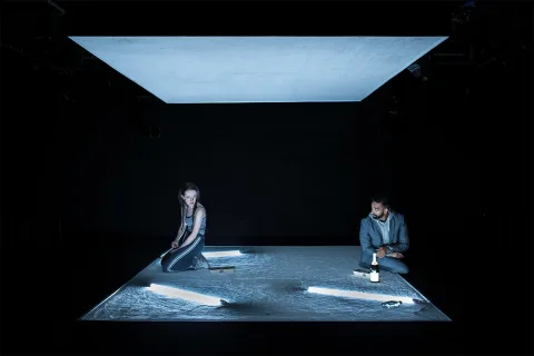 Tia (Mollie Milne) and AJ (Taqi Nazeer) sit on a sparse stage, a grey flat roof hangs above them.