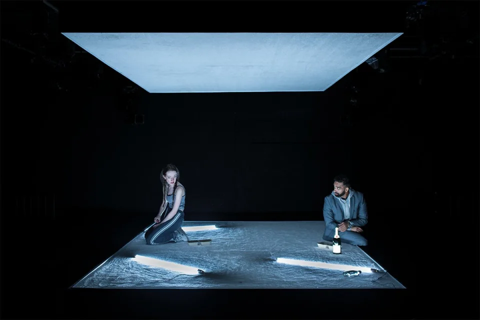 Tia (Mollie Milne) and AJ (Taqi Nazeer) sit on a sparse stage, a grey flat roof hangs above them.