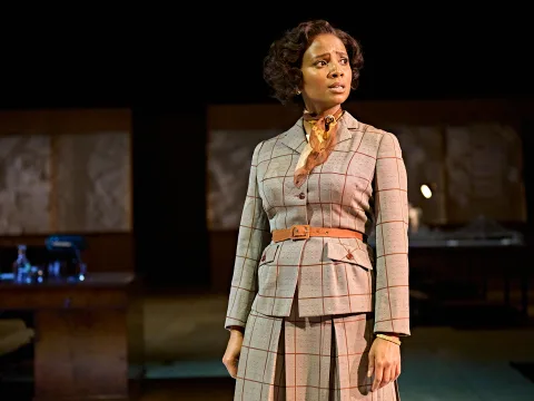 Alisha Bailey stands looking into the audience. She has a silk neck tie, short curls and pleated skirt.
