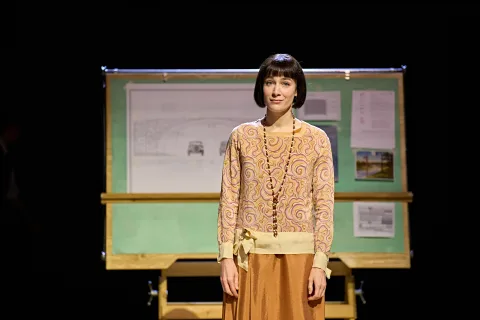 Siobhán Cullen stands in the middle of the image. She's wearing a sharp wig styles in a bob, a patterned thin jumper with a long beaded necklace. She looks pensive with a small smile.