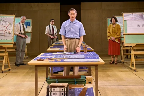 Samuel Barnett stands as the focus of the image in a blue shirt and brown smart trousers. He stands behind a big table with deign plans atop them. In the background are presentation boards with other architectural plans on.