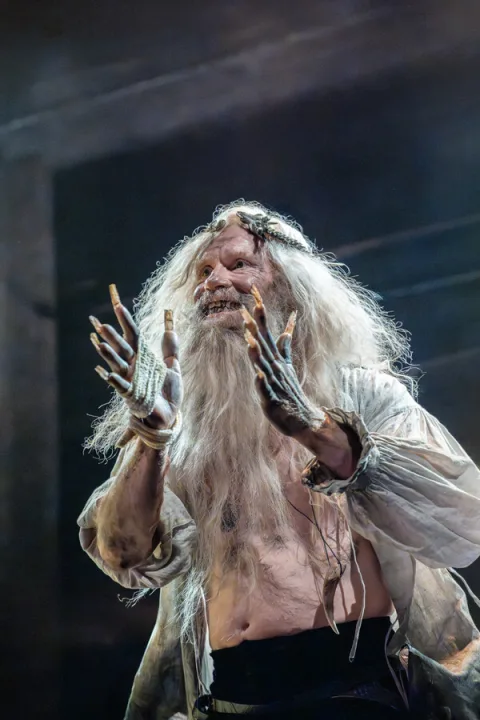 A man with a long straggly beard and hair and long grown out nails. He wears an unbuttoned dirty brown shirt and is smiling.