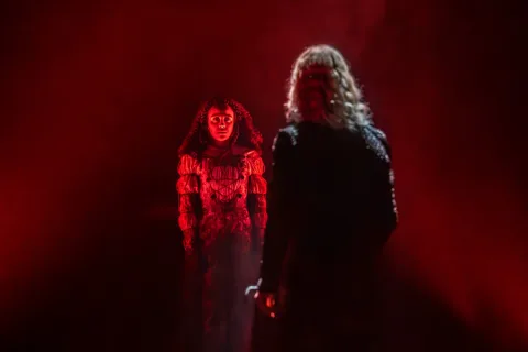 A young woman with curly black hair wearing an embroidered silver dress and shirt is illuminated by a red light. A man with blonde hair stands in the foreground opposite her.