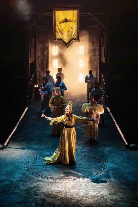 A woman stands in the centre of the image wearing an elaborate yellow dress and a cone shaped medieval hat on her head. Behind her are a crowd of people walking on to the stage and a large yellow banner above the stage.