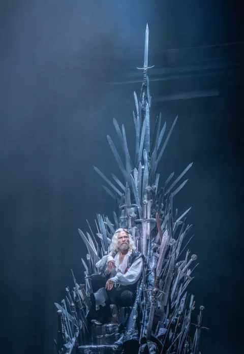 A man with long white hair sits in a large throne made of metal swords. We can see some smoke behind him.