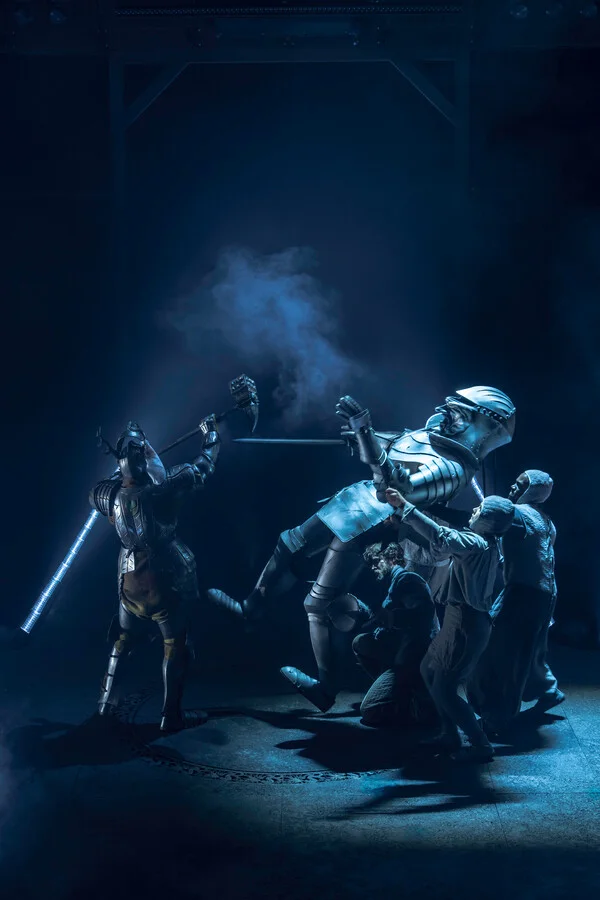 A large puppet soldier in a full suit of armour is being operated by a team of puppeteers. Stood opposite is a man in a full suit of armour lifting a battleaxe. Smoke is behind them.