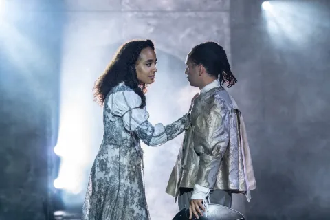 A man and woman stand opposite each other. The woman wears an embroidered silver dress, the man a white shirt and sliver jacket. The woman has her hand on the man’s chest.