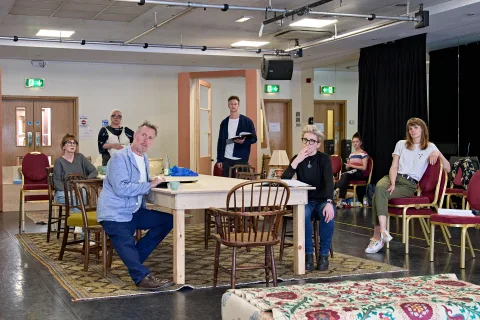 A large rehearsal room is filled with a big table atop a woven rug. There are different styles of wooden chairs sat at the table and also around the room. Seven people can be seen (two men and five women), most of them are sitting. They all look off in th from The Southbury Child