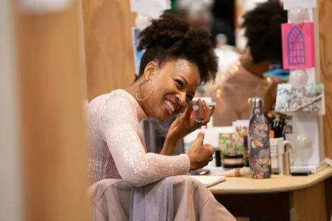 Racheal Ofori ( who has dark afro hair in a high ponytail, wearing huge hoop earrings and a sparkly pink top) is sat at a dressing table listening to her phone with a huge grin on her face