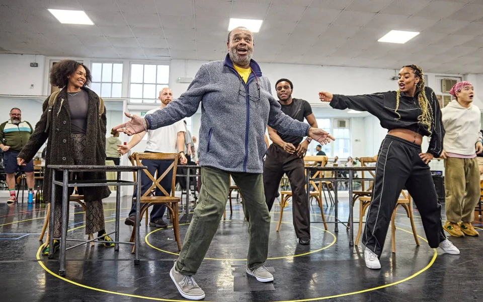 A group of people rehearse a lively musical number in a bright studio, with a man in a gray jacket standing center stage, arms open, and others dancing and smiling around him. Wooden chairs and tables are scattered nearby.