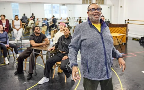A smiling man stands at the front of a dance studio while a group of seated people behind him watch. The room has mirrors, chairs, and a piano, with a lively and engaged atmosphere.