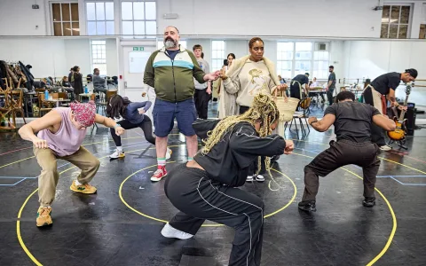 A group of people rehearsing in a spacious studio, some squatting and dancing energetically while others stand and observe. Musical instruments and chairs are scattered in the background.