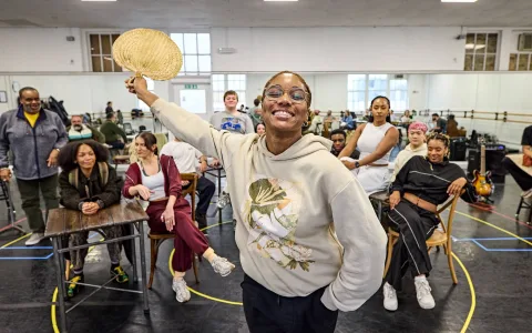 A smiling person in a light hoodie holds a woven fan up, standing in front of a diverse group of people sitting and standing in a spacious, mirrored rehearsal studio.