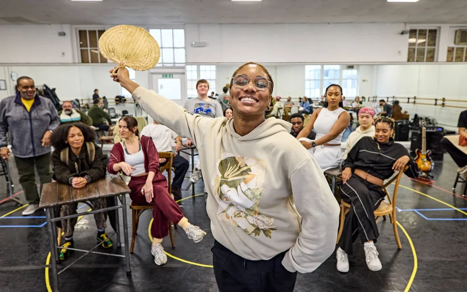 A smiling person in a light hoodie holds a woven fan up, standing in front of a diverse group of people sitting and standing in a spacious, mirrored rehearsal studio.